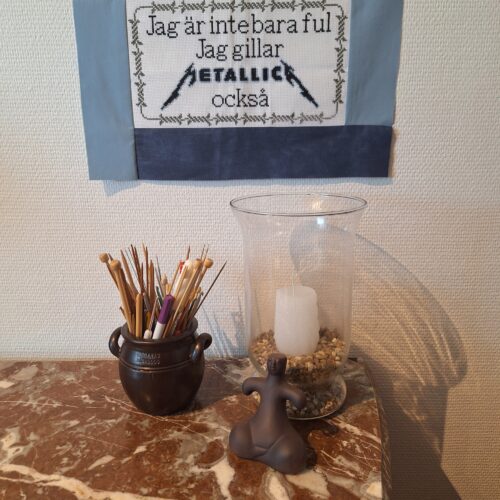 Ett korsstygn med texten "Jag är inte bara ful, jag gillar Metallica också" som är uppsatt på en vit vägg.